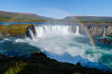 Fototapeta premium Experience the stunning beauty of Godafoss waterfall plunging into turquoise waters, surrounded by lush greenery and a vibrant rainbow under a clear blue sky, showcasing natures essence in Iceland
