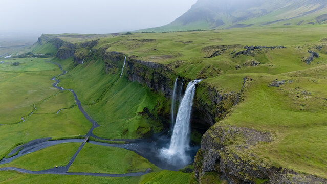 Seljalandsfoss waterfall flows dramatically over cliffs, surrounded by rolling green hills and a winding river. The misty atmosphere adds a magical touch to this iconic Icelandic landscape.