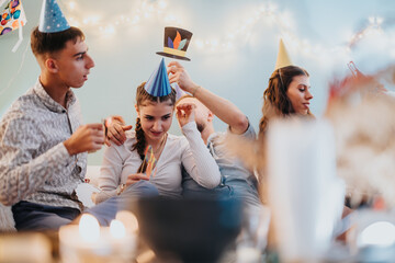 Young adults enjoying a cozy celebration at home, wearing party hats and sharing joyful moments...