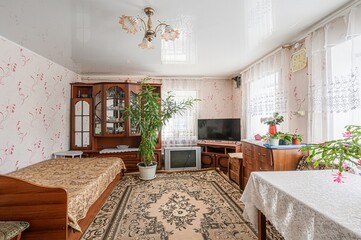 Cozy living room with a bed, wooden furniture, a large rug, and several houseplants. Floral...