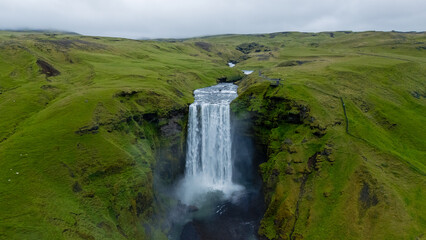 Skogafoss waterfall, set against a backdrop of lush green hills, exhibits its stunning cascade as water plunges dramatically. This natural wonder captivates visitors year-round.
