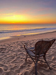 Wicker chair on sandy beach facing tranquil ocean waves at sunset, with vibrant colors illuminating...