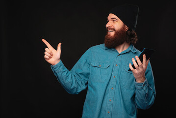 A joyful man in a denim shirt and hat pointing while holding a smartphone against a black...