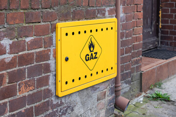 Bright yellow gas meter box displaying GAZ text and flame symbol mounted on an aged brick wall,...