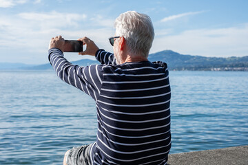 Rear view of a senior man sitting in front of the lake using his cell phone to take photographs, a...