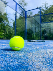 A bright yellow padel ball dominates the foreground with razor-sharp detail, while a professional racket lies slightly blurred on the blue court surface behind it. 