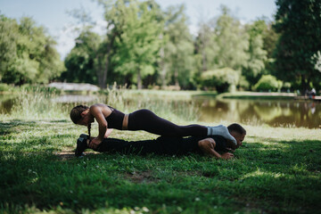 A couple engages in a physical workout on a sunny day in a park by a lake, showcasing teamwork,...