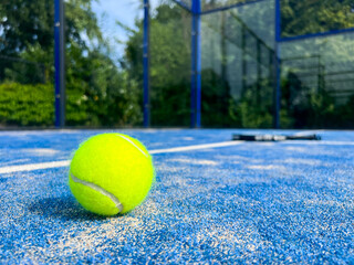 A bright yellow padel ball dominates the foreground with razor-sharp detail, while a professional racket lies slightly blurred on the blue court surface behind it. 