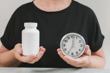 An elderly woman holds an alarm clock and a bottle of medicine.