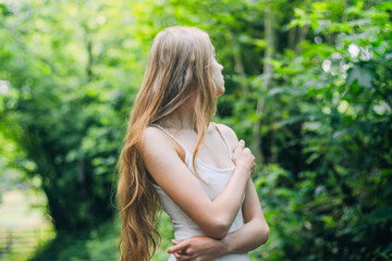 A young woman stands in a vibrant green forest, her long hair flowing freely as she looks away. The...