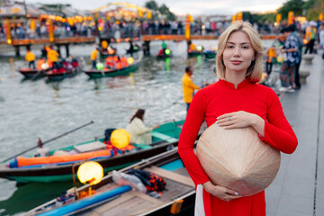 Female in traditional attire by riverside in hoi an, vietnam. River with tourist boats lantern lit