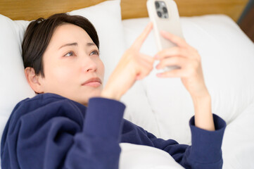 A Japanese woman lying on her back in bed looking at her smartphone and worrying