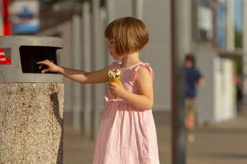 A little girl wearing a bright pink dress is happily throwing an ice cream cone into a nearby trash...
