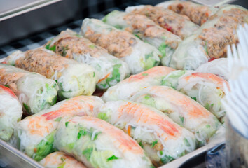 Close-up of fresh shrimp spring rolls filled with rice noodles, lettuce, and vegetables, wrapped in translucent rice paper. Displayed in a tray at a vibrant outdoor street food festival. 