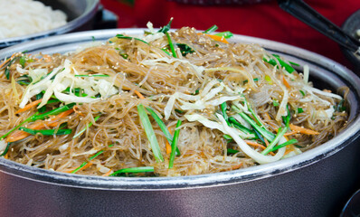 Close-up of traditional Asian glass noodles (funchoza) stir-fried with fresh vegetables such as carrots, cabbage, and green onions. The dish is served in a large metal pan at a street food market.