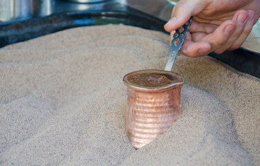 Close-up of a hand holding a cezve (copper coffee pot) brewing traditional Turkish coffee in hot...