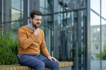 A young man wearing glasses sits on a bench near a building and suffers from a sore throat, holding...
