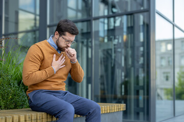 A young man is sitting on a bench outside, feeling unwell, holding his hand to his chest and...