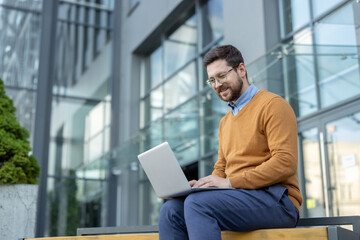 Smiling young man sitting on the street and using a laptop which he holds on his legs