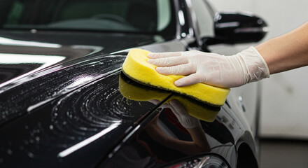 Man waxing black car surface with yellow sponge, focusing on detailing indoors