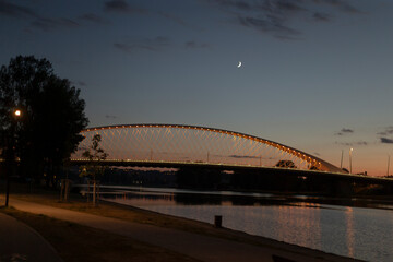 A magnificent bridge reflects in calm waters at dusk, framed by a crescent moon and soft clouds,...