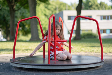 A five-year-old girl rides a carousel on a playground. A Caucasian girl in a red T-shirt walks on...
