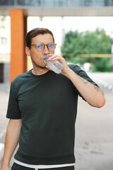 A young man in a dark green shirt and glasses drinks water from a clear bottle while standing in a...