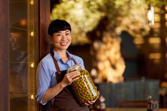 Smiling Woman Holding a Jar of Green Olives in a Rustic Interior Setting - Powered by Adobe