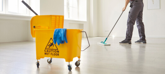 Janitor with mop cleaning empty office floor, worker mopping by yellow cart with professional...