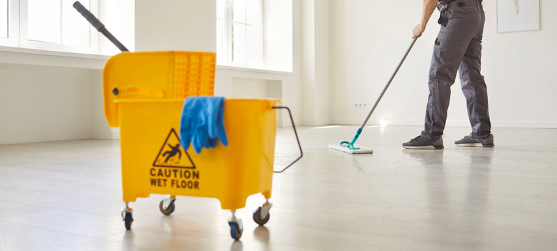 Janitor with mop cleaning empty office floor, worker mopping by yellow cart with professional cleaners supplies and warning wet floor sign to clean room. Cleanup service, sanitation, hygiene concept - Powered by Adobe