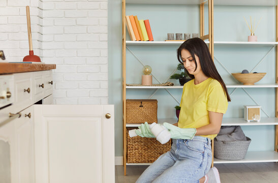 Woman in rubber gloves kneels on floor in kitchen, holding broken pipe with concerned or frustrated expression. Plumbing issue, unexpected household emergency, need for professional repair services