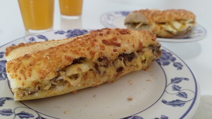 Close-up of a delicious toasted baguette sandwich with melted cheese and meat filling, served on a classic blue-patterned plate. In the background, glasses of orange juice complete the meal.