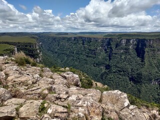 Scenic view from Canion Fortaleza, a rocky cliff edge overlooking a deep, lush green canyon filled with dense forest under a dramatic blue sky. A scenic natural landscape of a grand valley and plateau