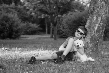 A boy sits under a tree with his Maltipoo puppy. A boy walks with a beautiful white Maltipoo puppy