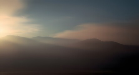 Obraz premium Atmospheric landscape featuring soft mountains shrouded in mist illuminated by a gentle sun creating hazy beams of light. The sky transitions from a muted blue to warmer hues.