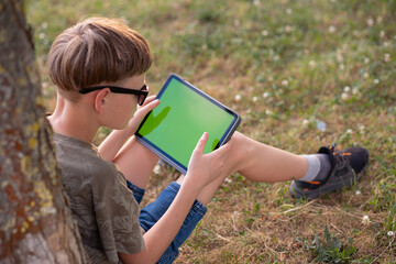 A boy sits in a park under a tree and plays on a tablet. The boy holds a tablet in his hands. Gadget addiction
