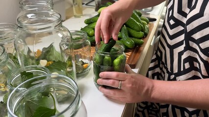 A woman prepares homemade pickles, placing cucumbers into jars with herbs, highlighting DIY crafts and culinary traditions
