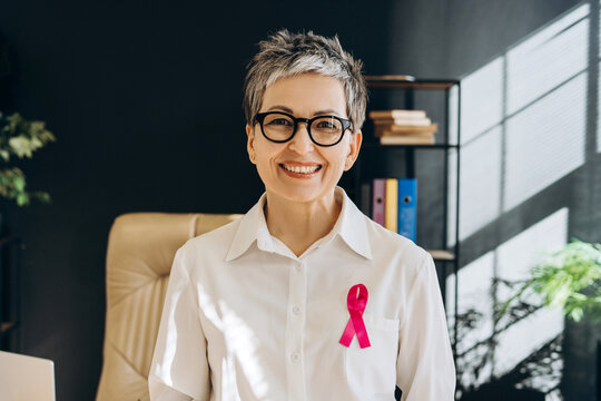 Smiling senior professional woman wearing pink ribbon supporting breast cancer awareness - Powered by Adobe