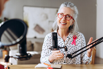 Senior woman with pink ribbon talking on microphone recording podcast about breast cancer awareness