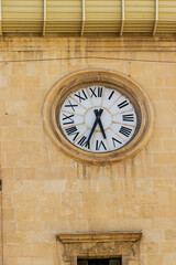 Fototapeta premium Close-up of a large clock with Roman numerals on a stone wall, showcasing intricate detailing and classic design. The clock is an architectural feature in Alicante, Spain.