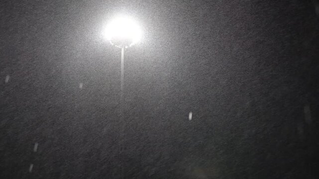 A street light is lit up in the dark. The sky is cloudy and the snow is falling