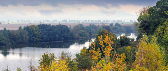 A river winding through autumn trees and fields, with a cloudy sky, creating a calm and picturesque natural landscape