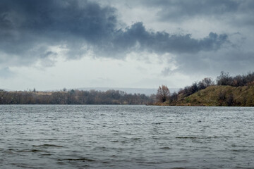 Dark rainy sky over a river with a tree line on the horizon, creating a gloomy yet peaceful natural...