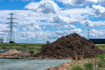 Construction site with large dirt mound, power lines, and pylons under partly cloudy blue sky in...