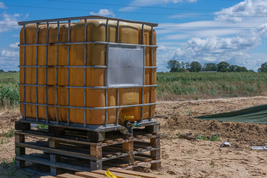 Yellow IBC water tank on wooden pallets at construction site with sandy ground and green field in background.