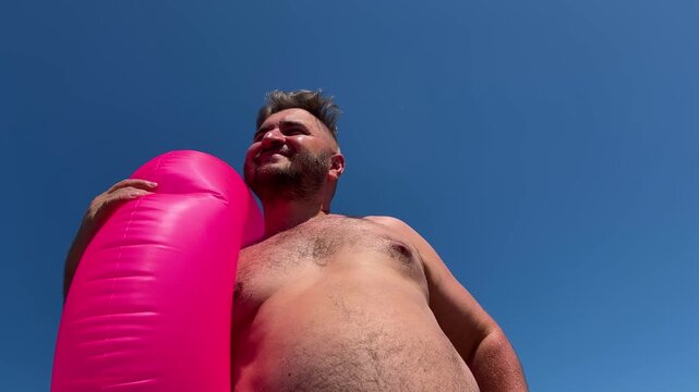 Horizontal 4K video. Bottom view. Fat mature white man with beard, big belly, pink swim ring on sea beach put hand to forehead to shield eyes from sun shine. Sun day. Blue sky. Travel, tourism, trip