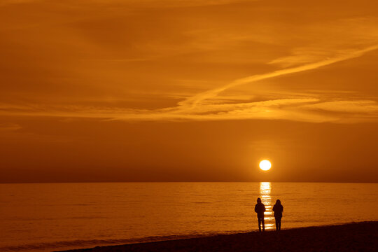 back rear view of hugging couple stands on pebbled coast beach and enjoys the intense sunset over the Black sea in Batumi. pair wearing warm jackets. Autumn spring season. Seasonal image.