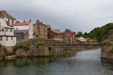 Fototapeta premium An outdoor shot showcases a body of water with buildings on the bank. The buildings are various colors and styles with a bridge over the water in Staithes - North Yorkshire - United Kingdom