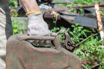 A weathered, rusty gas can is held by a gloved hand against a natural outdoor backdrop