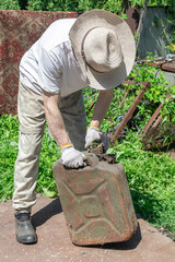 A person lifts an old, rusty gas can while wearing a hat and gloves outdoors.